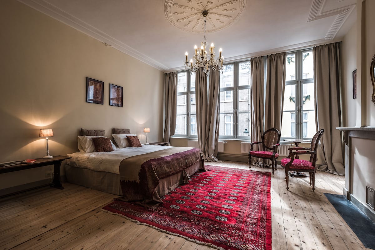 A bedroom with a large bed, a chandelier, a red patterned rug, and a fireplace.