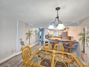 A dining area with a glass-top table and wicker chairs, a kitchen with white cabinets, and a ceiling fan.
