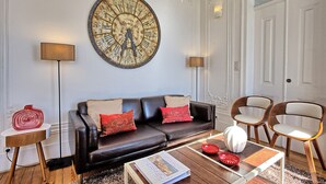 Un salon avec un canapé en cuir noir, des coussins rouges et beige, une horloge murale ronde, deux lampadaires, une table basse en bois ornée d’assiettes décoratives rouges et un vase blanc.