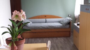 Una habitación pequeña con una mesa de madera, una cama individual con colcha a cuadros, una planta en maceta y un área de cocina pequeña.