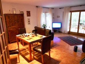 Ein Wohnzimmer mit einem Holzesstisch, Stühlen, einem Fernseher und einem großen Fenster mit Vorhängen.