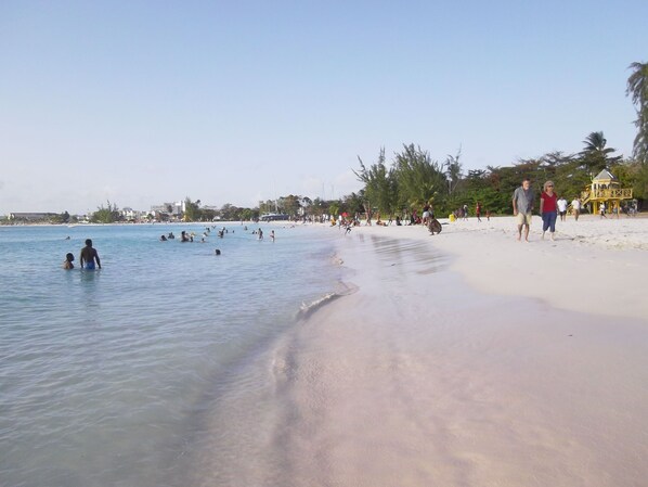 A beach with clear water, people swimming and walking on the sand, and a lifeguard tower in the background.