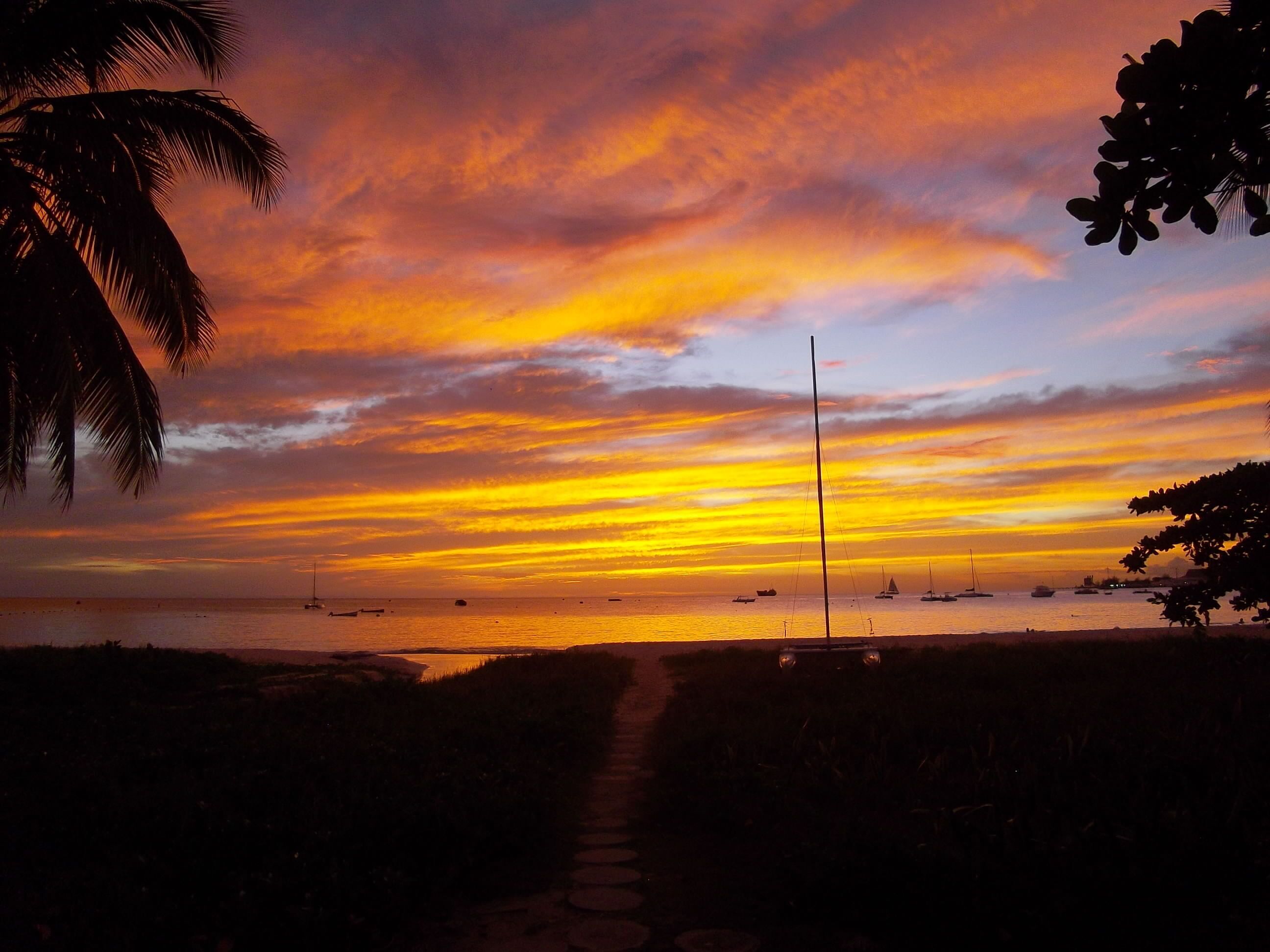 Una vista al atardecer con un camino que lleva al agua, barcos y un velero a lo lejos.