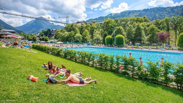 Un espace piscine où des personnes se prélassent au soleil sur la pelouse, avec une chaîne de montagnes en arrière-plan et un téléphérique visible au loin.