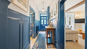 A hallway with blue walls, a wooden bench, a floral rug, and a door leading to a room with a white couch and a table with oranges.
