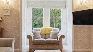 A cozy living room with a sofa facing a large window, a flat-screen TV mounted on the wall, and a decorative wall with floral wallpaper.