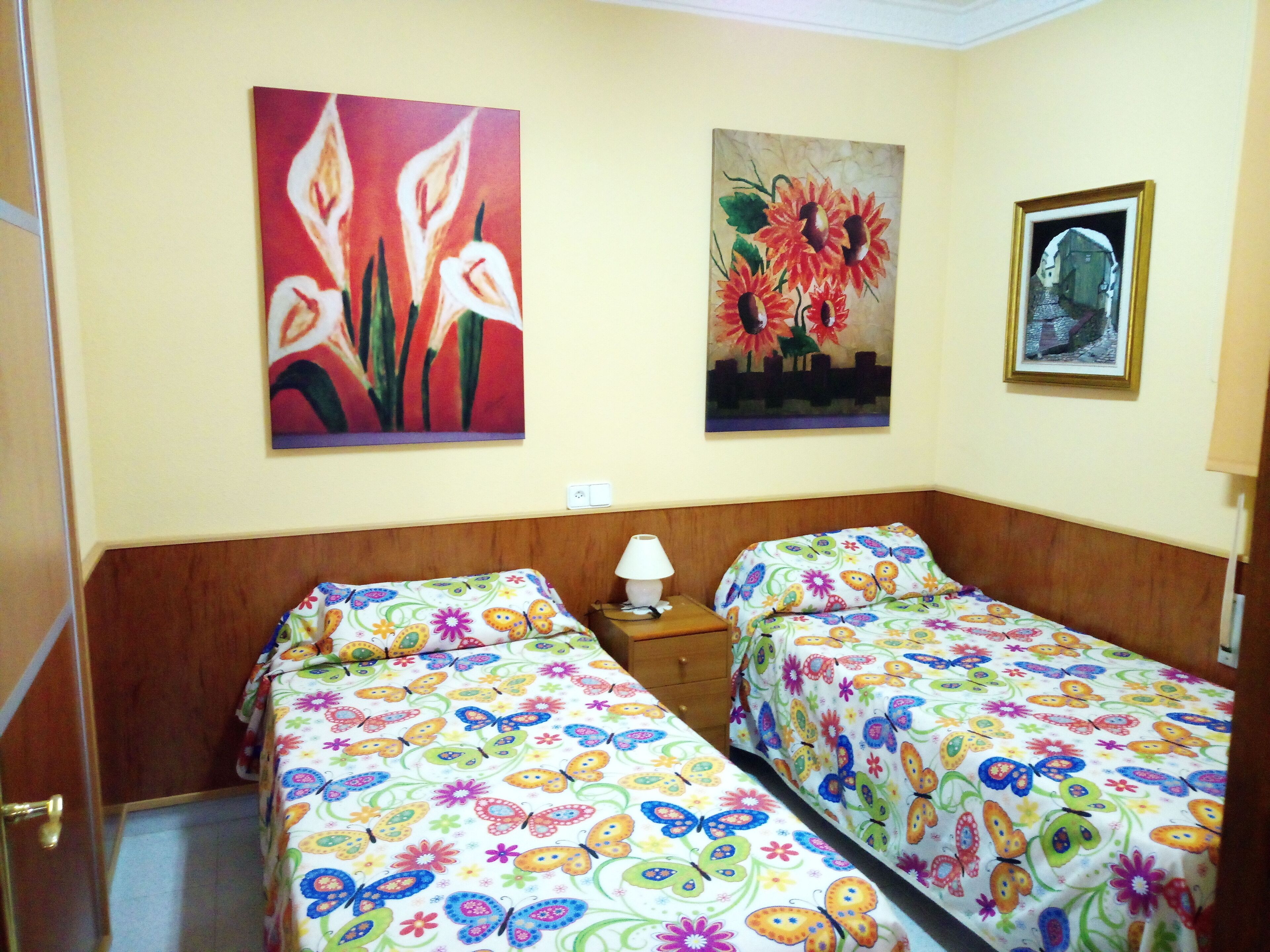 A room with two beds, each with a colorful butterfly and flower bedspread. There are two paintings on the wall, one depicting flowers and the other a cityscape.