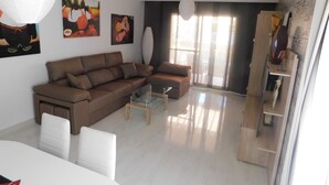 A modern living room with a brown sectional sofa, a glass coffee table, and a white dining table with chairs. There are abstract paintings on the wall and a large window with curtains.