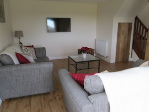 A living room with a grey sofa, a small table with a potted plant, and a TV on the wall. - Barn conversion in beautiful, peaceful, rural location with stunning views. (Hailsham)