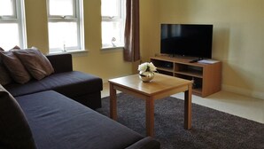 A living room with a sofa, a coffee table with a vase, a TV on a wooden stand, and windows with curtains.