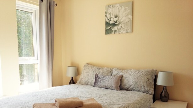 A bedroom with a bed, two bedside tables, a window with curtains, and a wall-mounted artwork.