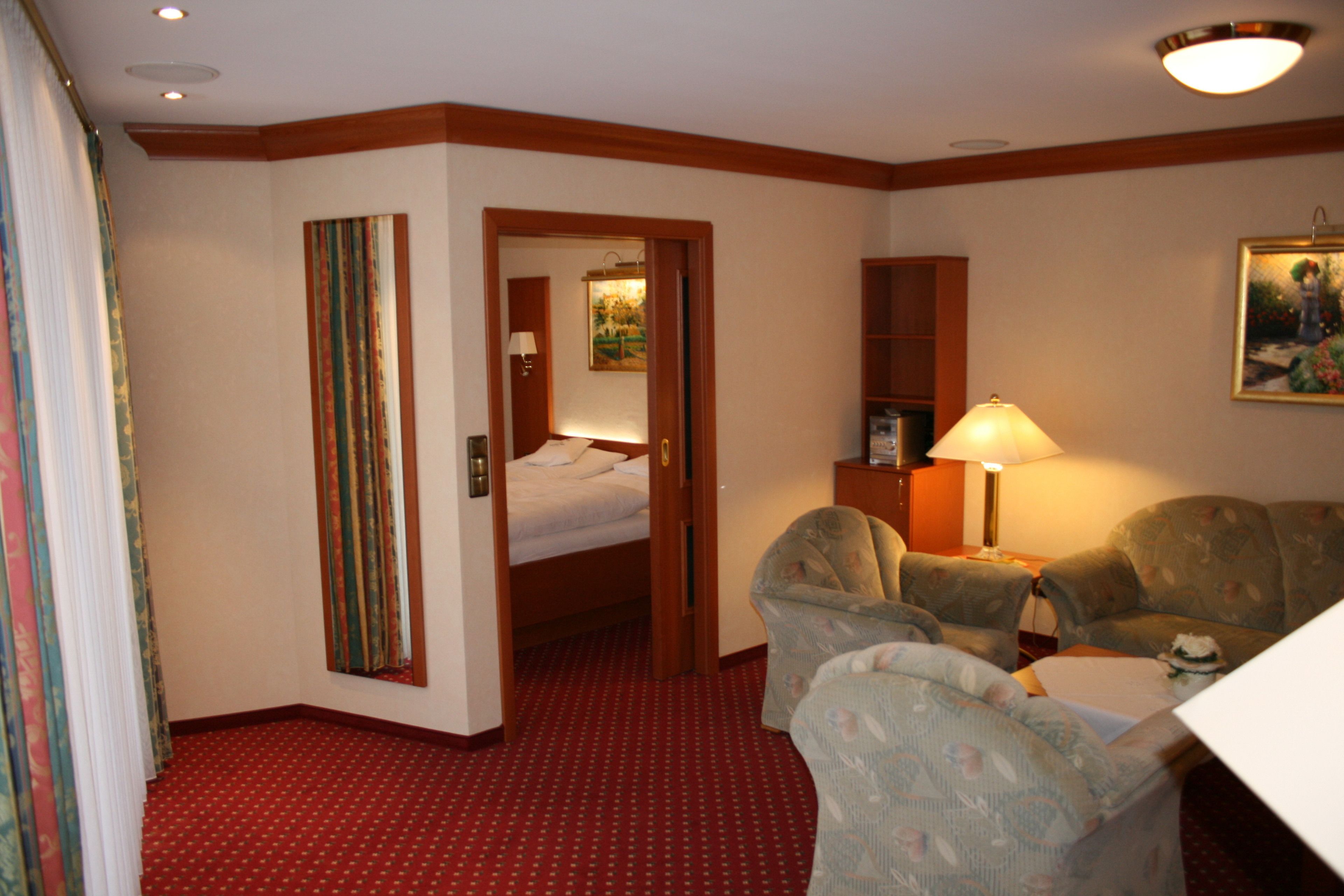 A hotel room with a bed, a desk, a chair, a lamp, a painting, and a red carpet.