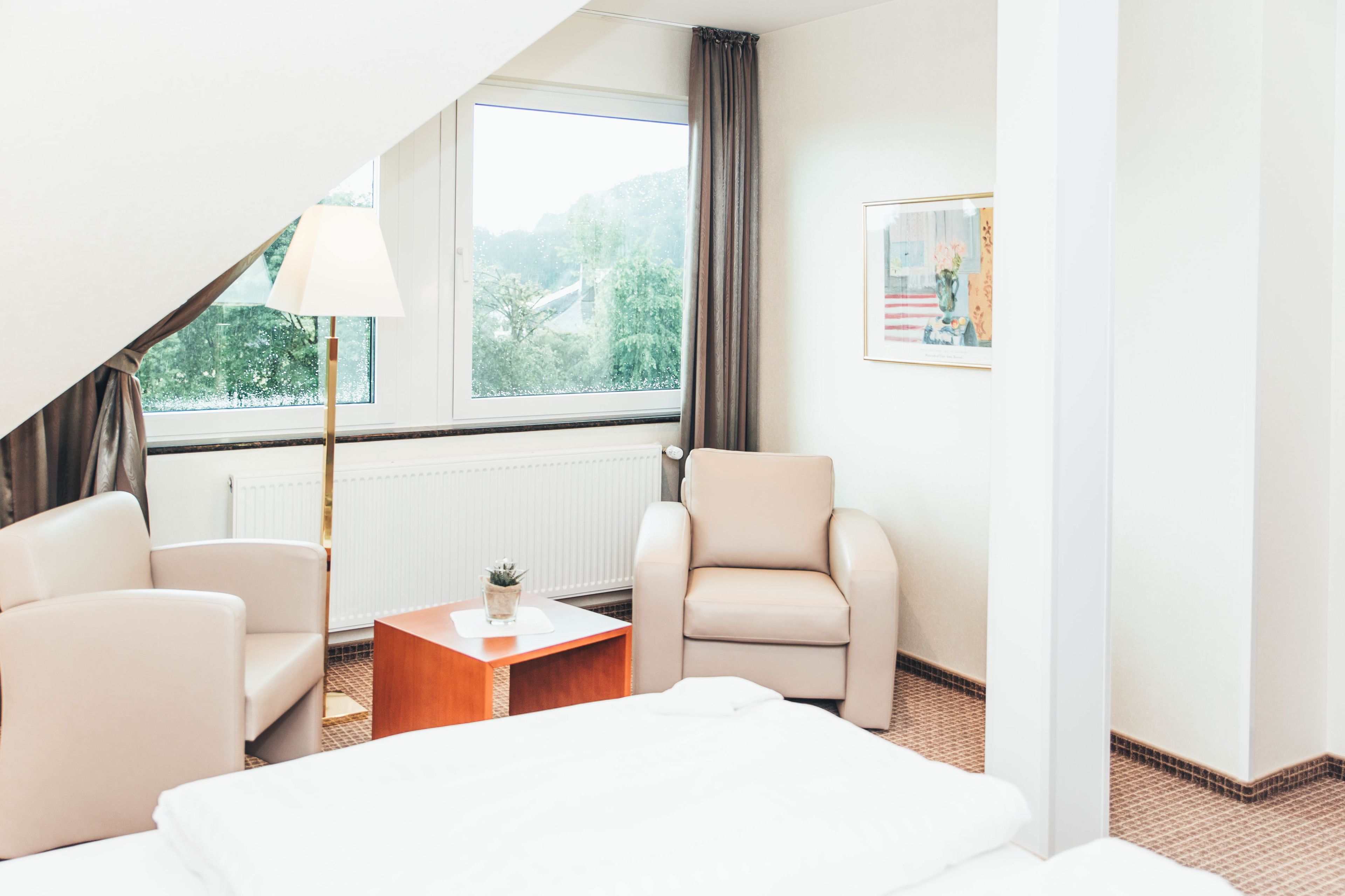 Une chambre d’hôtel avec un lit, deux fauteuils, une petite table avec une lampe, un tableau au mur et une fenêtre donnant sur un paysage verdoyant.