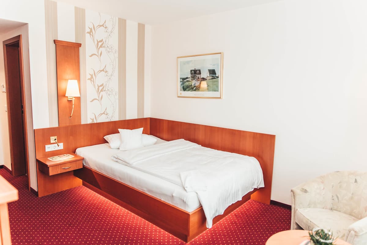 Une chambre d’hôtel avec un lit en bois, une table de chevet, une lampe, un tableau encadré au mur et un sol recouvert d’une moquette rouge.
