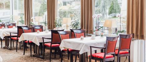 A dining area with tables set for four, each with a white tablecloth and red cushioned chairs.