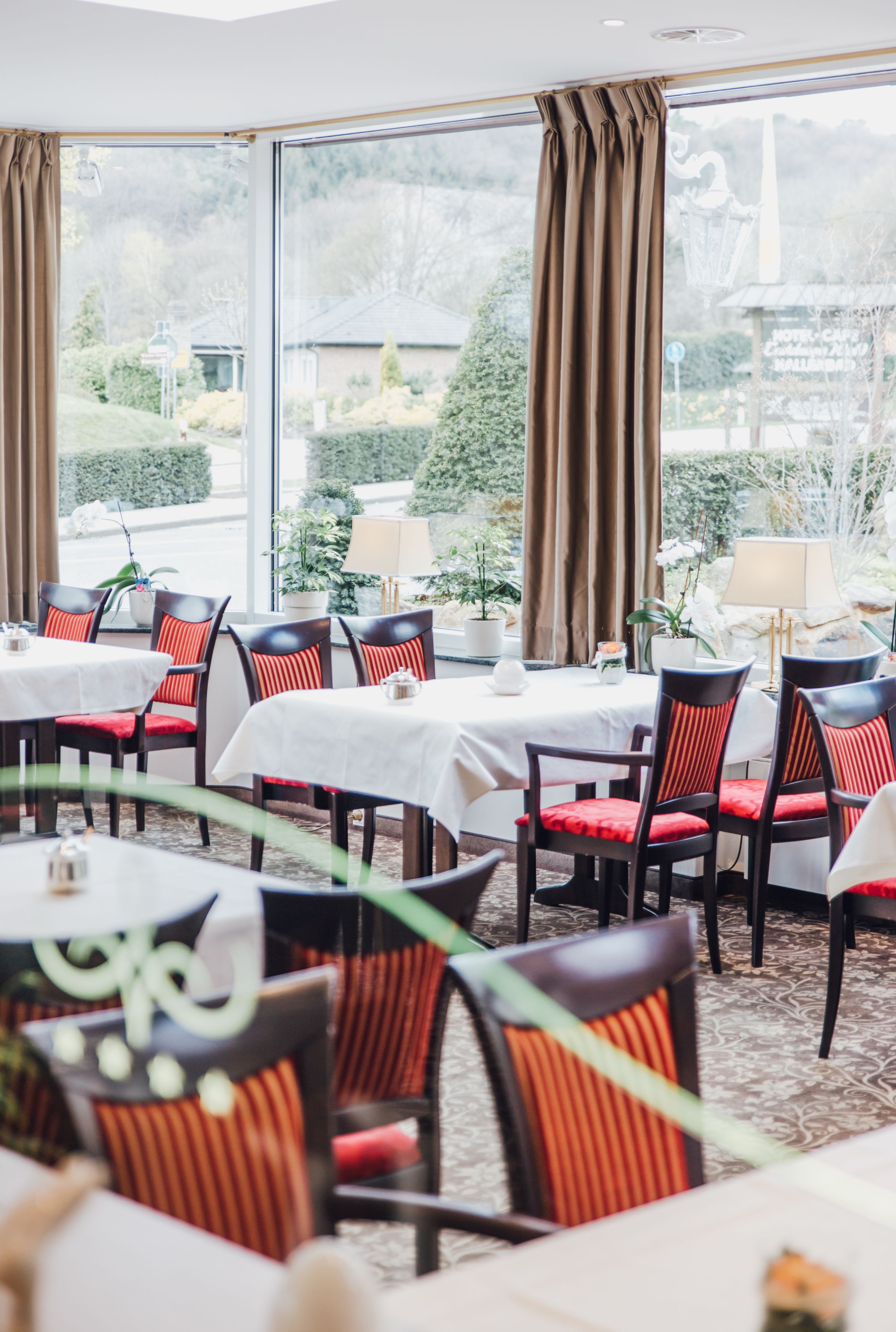 Un restaurant avec des tables et des chaises, de grandes fenêtres avec des rideaux, et une vue sur la verdure et les bâtiments environnants.