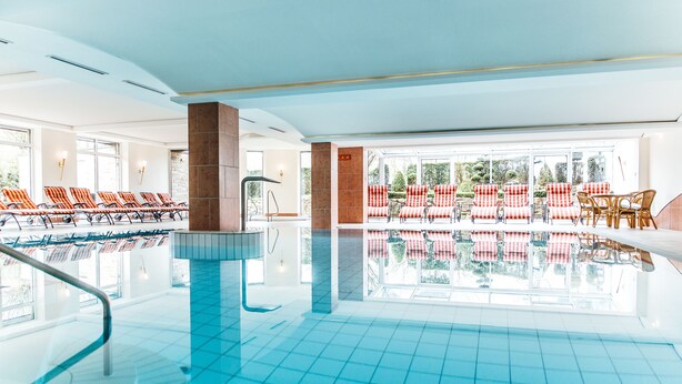 An indoor swimming pool with tiled flooring, red and white checkered lounge chairs, and a glass-enclosed area with more chairs and a table. - Ringhotel Teutoburger Wald (Tecklenburg)