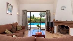 Un salon comprenant un canapé, une table basse avec un livre et un vase, un téléviseur, une cheminée, et une vue sur une piscine et un espace verdoyant.