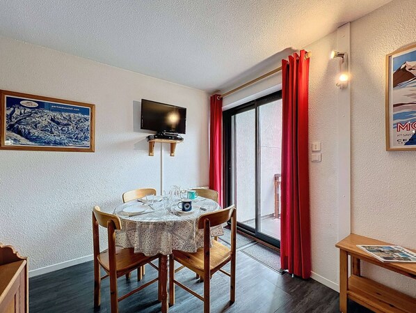 Un comedor con una mesa dispuesta para dos, un televisor montado en la pared y un ventanal con cortinas rojas.