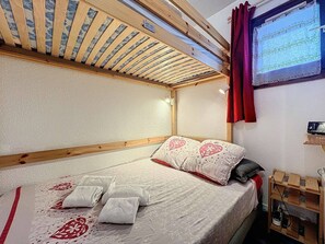 Un dormitorio con cama, almohadas, cabecero de madera, televisor montado en la pared y una pequeña estantería de madera.