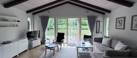 A spacious living room with a high ceiling, a flat-screen TV, a sofa, a coffee table, and a large window offering a view of the outdoors.