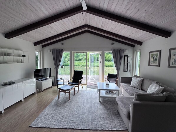 A spacious living room with a high ceiling, a flat-screen TV, a sofa, a coffee table, and a large window offering a view of the outdoors.