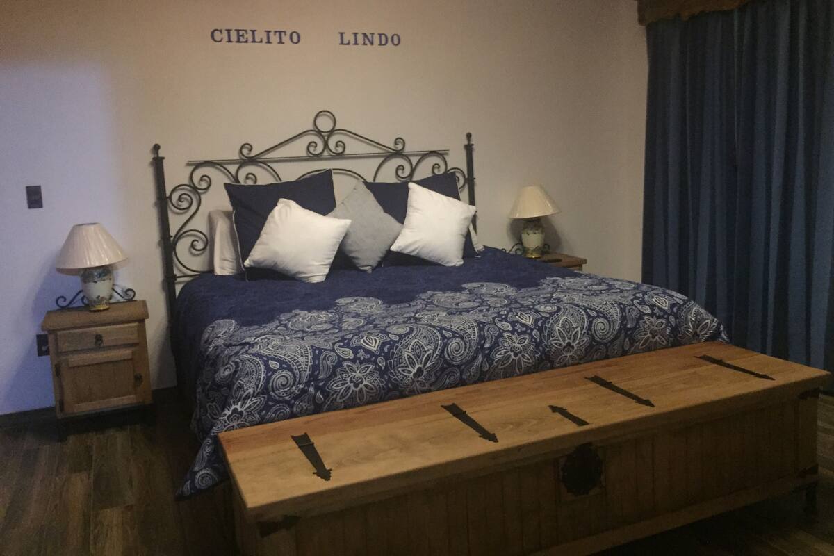 A bedroom with a metal bed frame, a wooden bench, two bedside tables with lamps, and a wall with the text "Cielito Lindo".