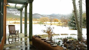 Un porche cubierto con una silla, un repisa de piedra con rocas y plantas, y vista a un paisaje nevado con árboles y montañas al fondo.