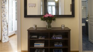 Een gang met een houten vloer, een donkere console tafel met boeken en een bloevas, een grote spiegel en een kroonluchter.