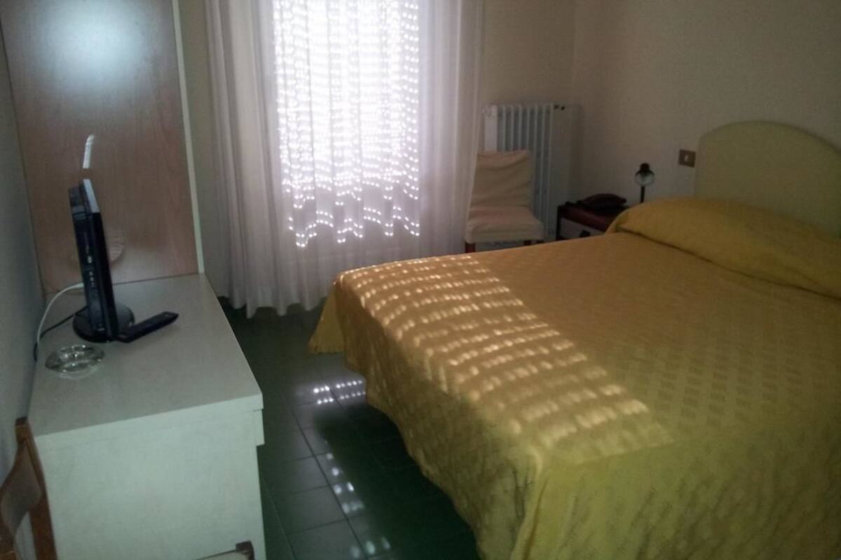 A hotel room with a bed, a desk with a computer, a chair, and a radiator.