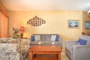 A living room with a grey sofa, patterned armchair, and a wooden coffee table with two martini glasses. A wall-mounted light fixture and a framed painting are visible.