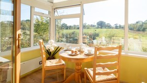 Een zonnige kamer met een gedekte theetafel, twee stoelen en uitzicht op een groen landschap door grote ramen.