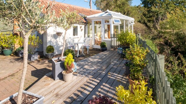 Een overdekt terras met een houten dek, potplanten en een witte pergola met glazen deuren.