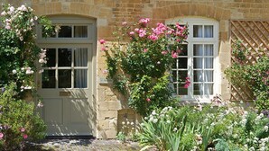 A stone building with two windows, one with a white frame and the other with a wooden lattice. There are pink roses and other flowers growing around the windows. - Bruern Cottages - Aintree (sleeps 6 + 1) (Chipping Norton)