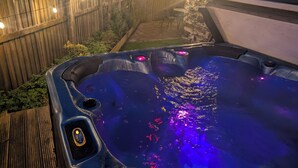 Un jacuzzi con panel de control, rodeado por una valla de madera y un muro de piedra, con luces colgantes al fondo.