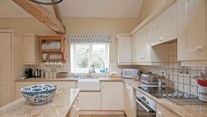 A kitchen with white cabinets, a wooden shelf with plates, a window with a valance, and a bowl on the counter. - Bruern Cottages - Shipton (sleeps 6 + 2) (Chipping Norton)