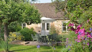 A stone building with a tiled roof, a garden with a table and chairs, and flowering plants. - Bruern Cottages - Shipton (sleeps 6 + 2) (Chipping Norton)