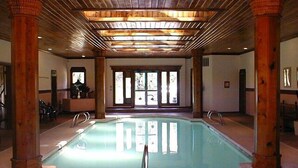 An indoor swimming pool with wooden beams and columns, a tiled floor, and a view of the outside through large windows.