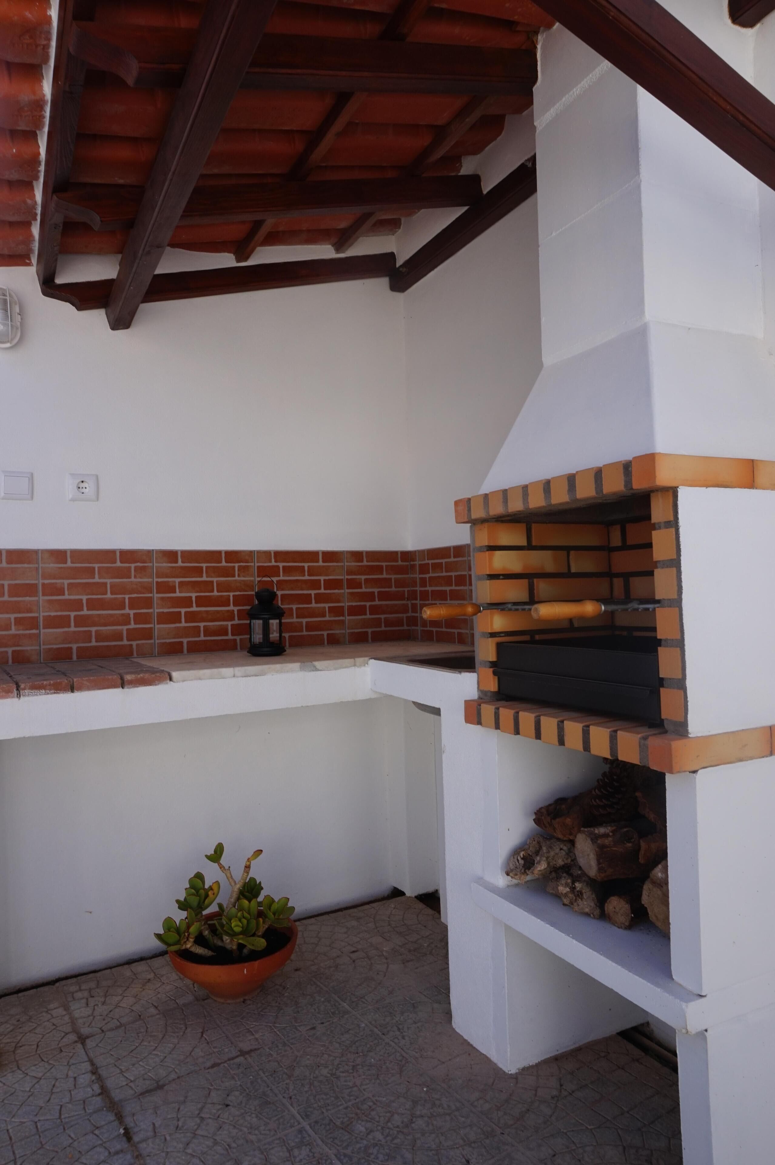 A brick oven barbecue area with a tiled backsplash, a potted plant, and a stack of firewood.