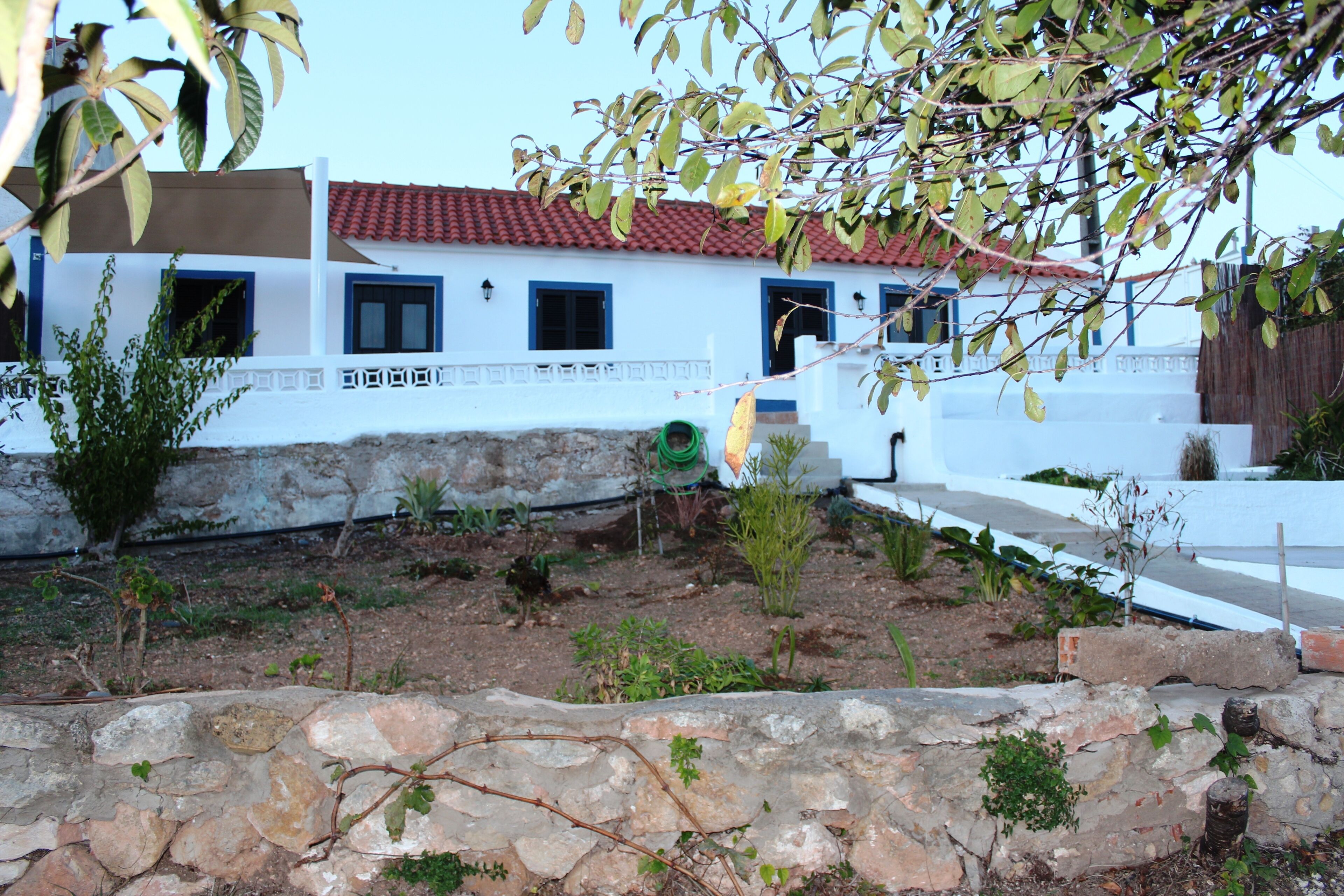 A white building with a red-tiled roof, a stone wall in the foreground, and a garden area with various plants.