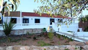 Un edificio blanco con techo de tejas rojas, un muro de piedra en primer plano y un jardín con diversas plantas.