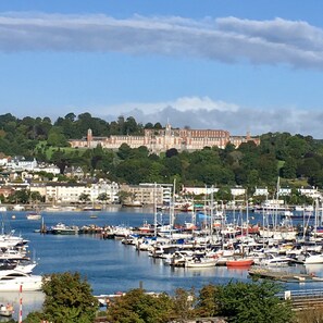 A marina with various boats docked, a large building with a tower in the background, and a hilly landscape with greenery.