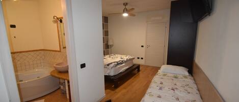 Un dormitorio con cama, mesita de noche, ventilador de techo y baño con bañera y lavamanos.