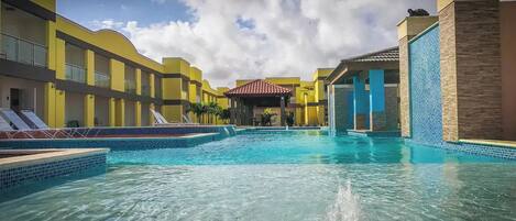 A swimming pool with tiled edges and a central fountain, surrounded by yellow buildings with balconies and a covered seating area.
