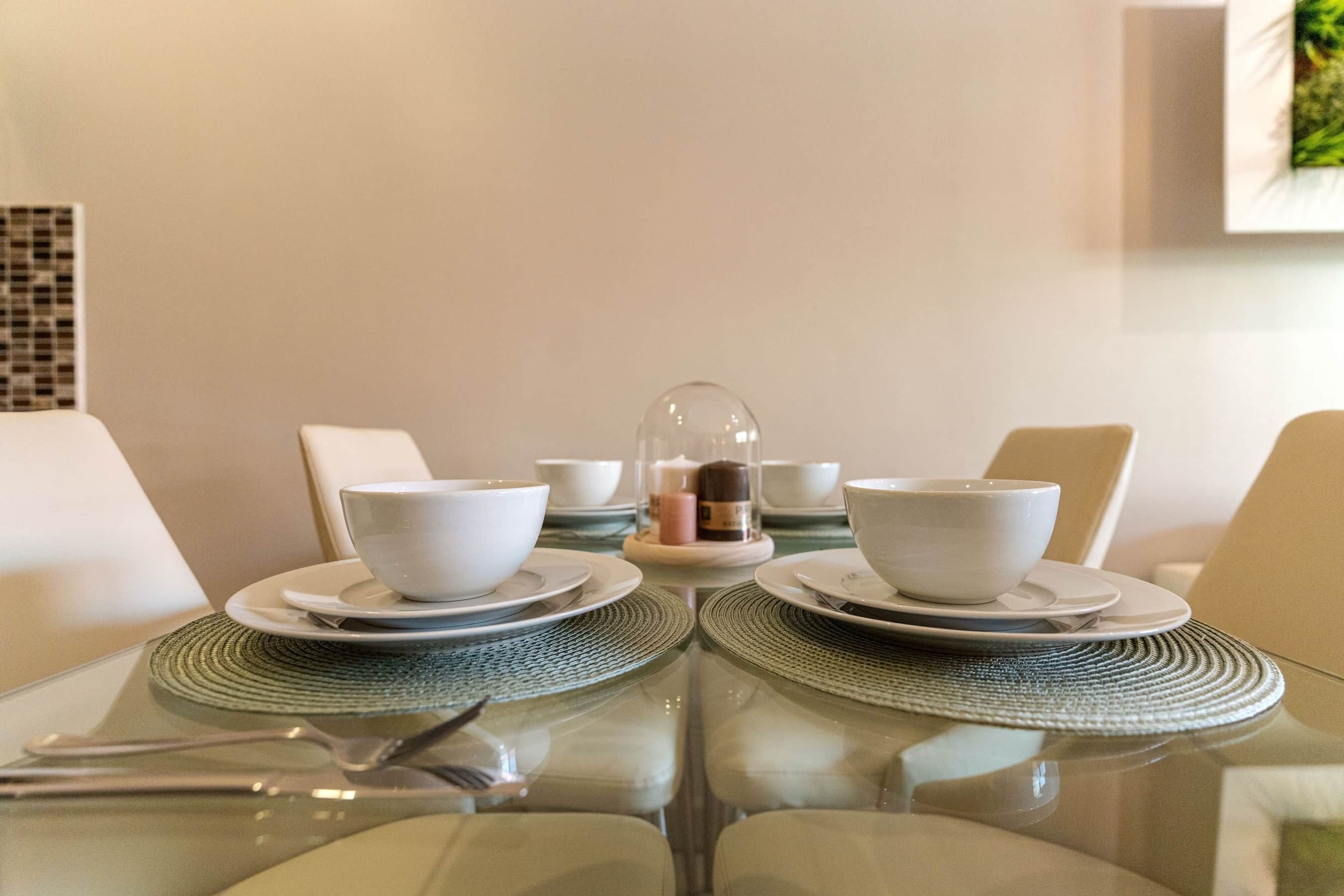 Une table à manger avec de l’assiette, des bols et des couverts blancs, le tout disposé sur un set de table en fibres tressées.
