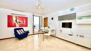 Un salon moderne avec un canapé bleu, une table à manger avec des chaises, un téléviseur fixé au mur et une grande fenêtre.