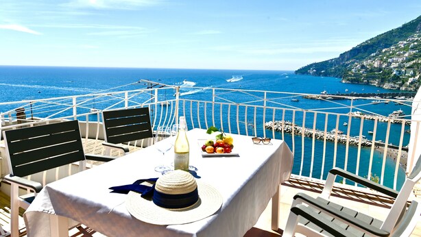 Une table est dressée sur un balcon qui offre une vue sur la côte, avec des bateaux, une côte rocheuse et des bâtiments situés sur une colline.