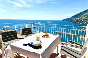 Une table est dressée sur un balcon qui offre une vue sur la côte, avec des bateaux, une côte rocheuse et des bâtiments situés sur une colline.