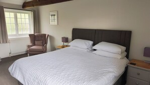 A neatly made bed with white bedding, a wooden nightstand with a lamp, a chair, and a window with curtains. - Wheat cottage, Chiddingly, spacious and well equipped character accommodation (chiddingly)