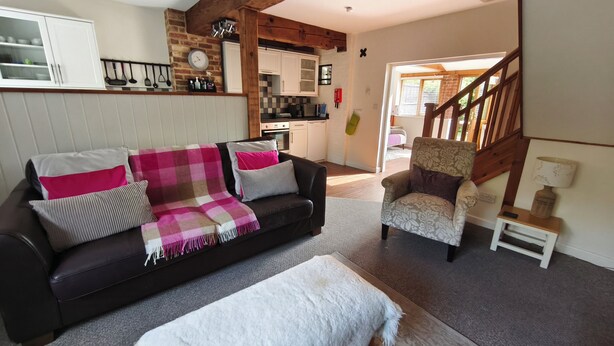A living room with a dark brown sofa, a white ottoman, and a patterned armchair. A kitchen is visible in the background with white cabinets and a brick wall. - Wheat cottage, Chiddingly, spacious and well equipped character accommodation (chiddingly)